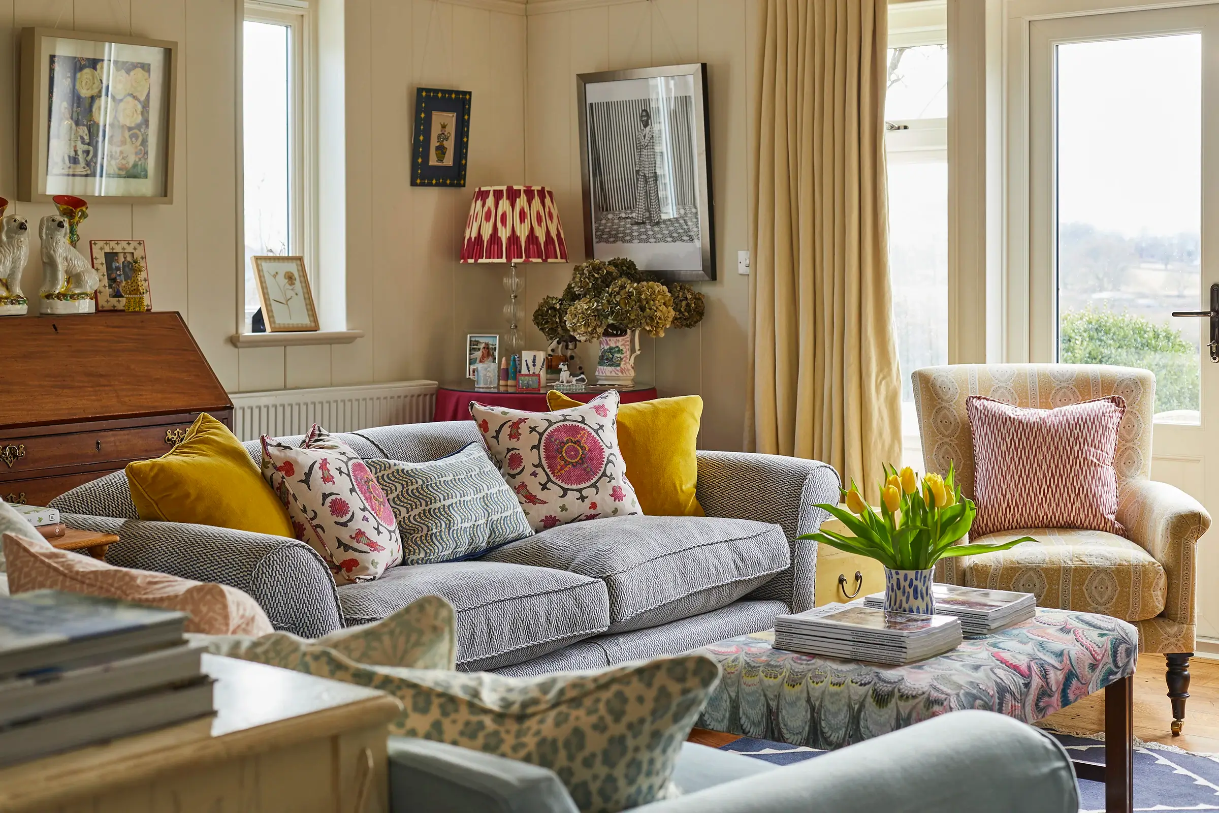 Cozy living room with a patterned sofa, colorful cushions, a floral lampshade, and a vase of tulips on the table.