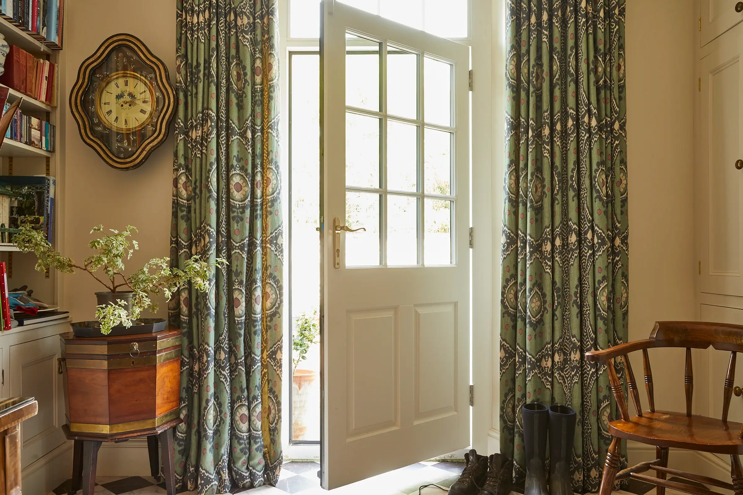 A door opens to a bright exterior, with patterned curtains, a clock, boots, and a wooden chair in the room.