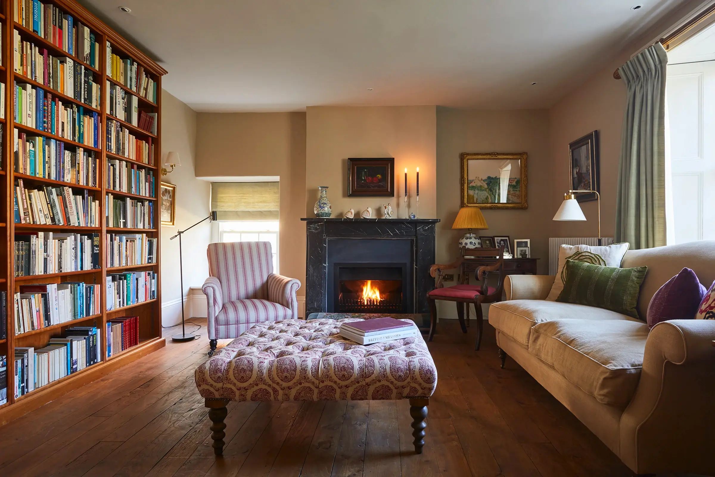 Cozy living room with bookshelves, armchair, tufted ottoman, and lit fireplace on a wooden floor.