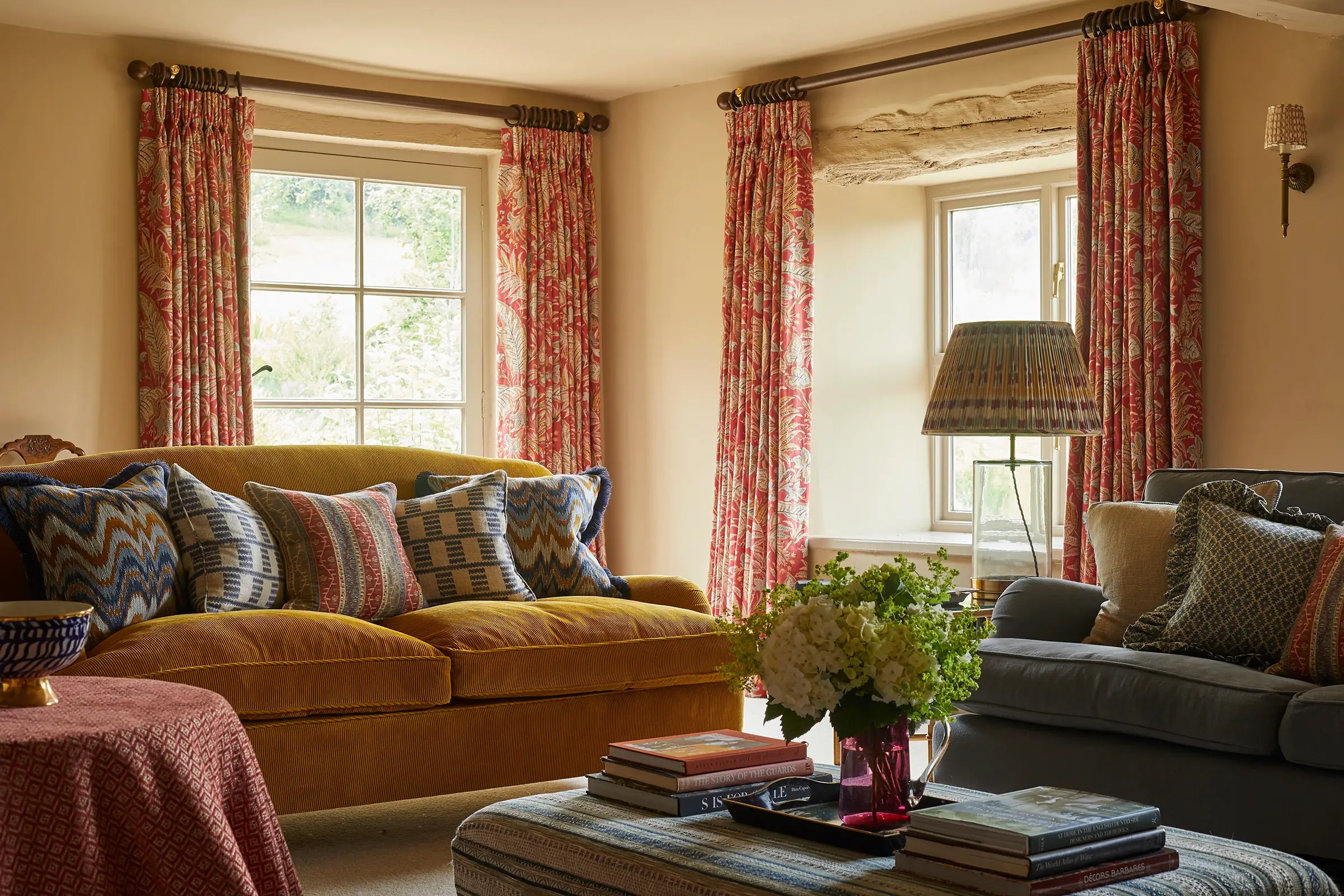 Cozy living room with couches, patterned pillows, floral curtains, table with books, and a potted plant.