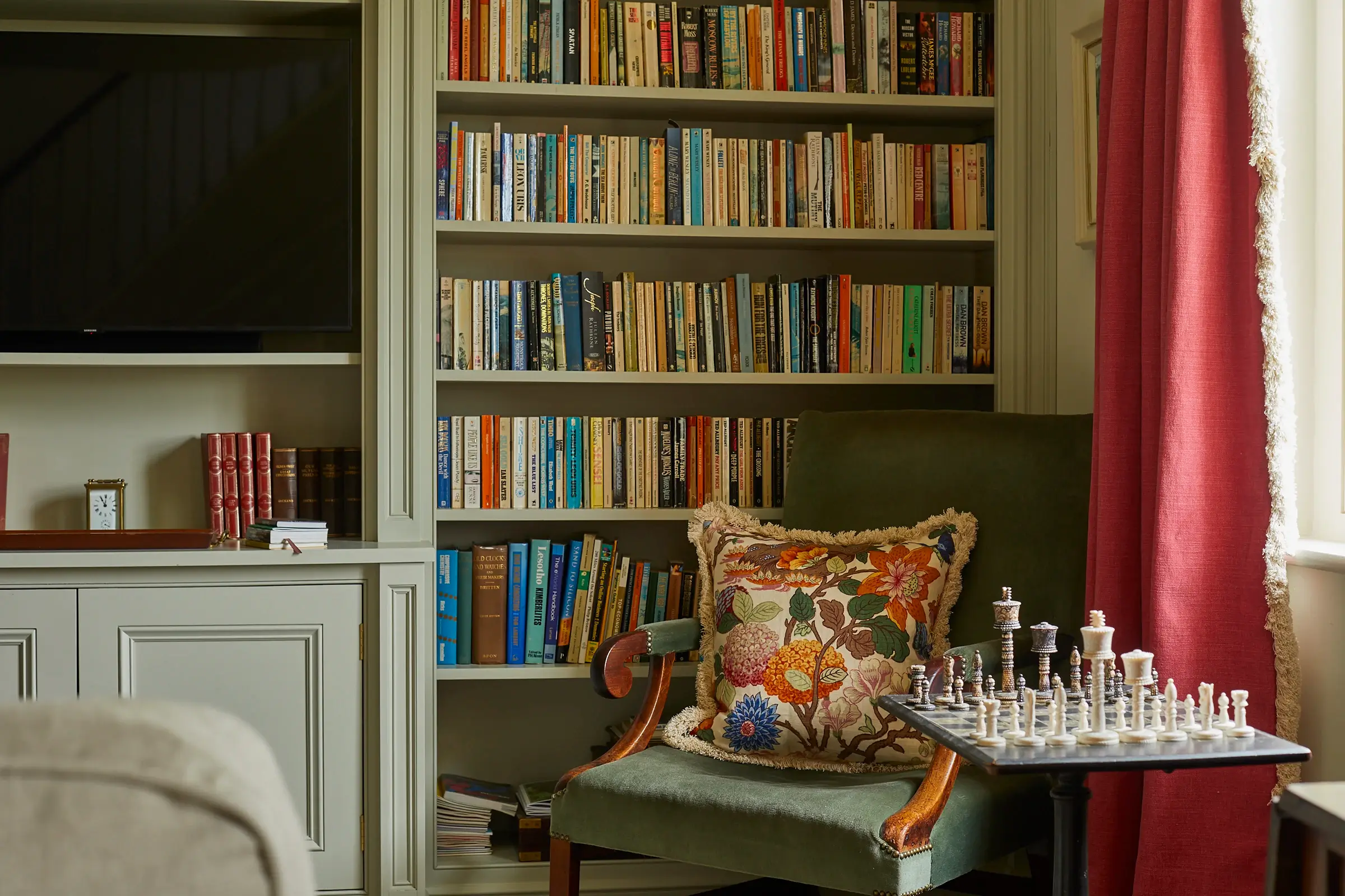 A cozy reading nook with a floral cushion, chess set, and a bookshelf filled with books in the background.