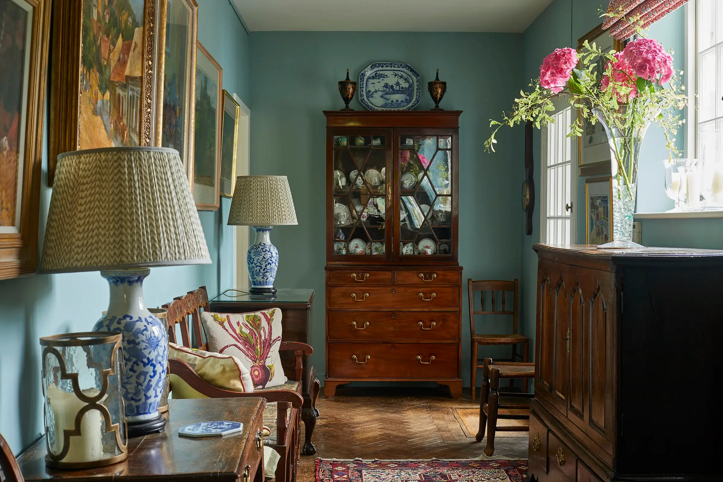 A cozy room with vintage furniture, including a wooden cabinet, lamps, paintings, and flowers on a table.