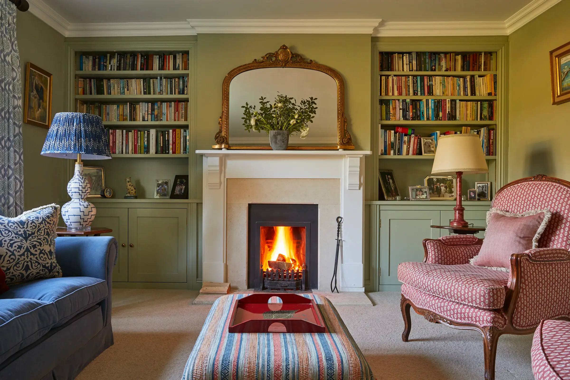 Cozy living room with a lit fireplace, bookshelves, blue sofa, patterned armchair, and a decorative mirror above the mantel.