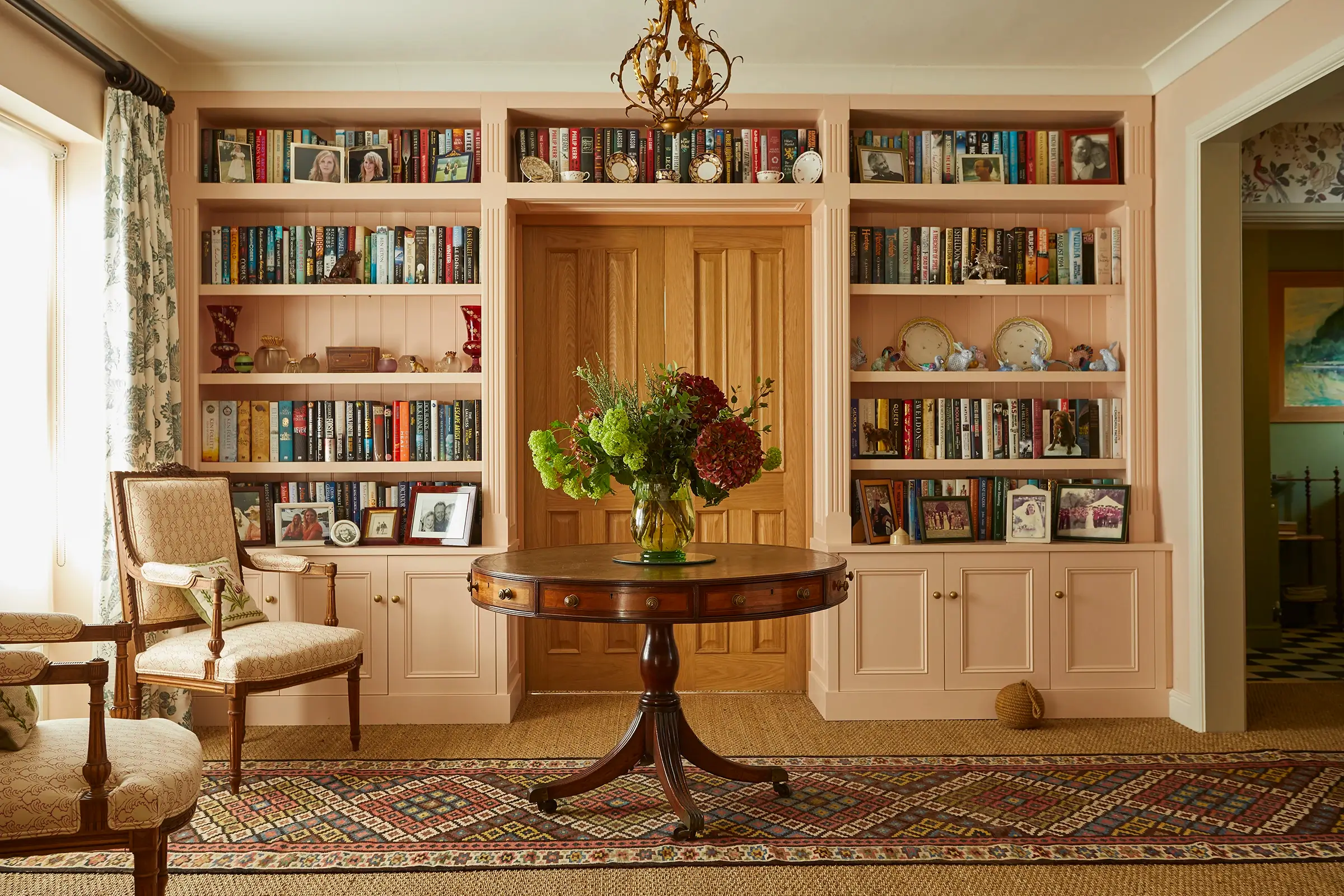 A home library with bookshelves, a round table with flowers, two chairs, and a chandelier overhead.