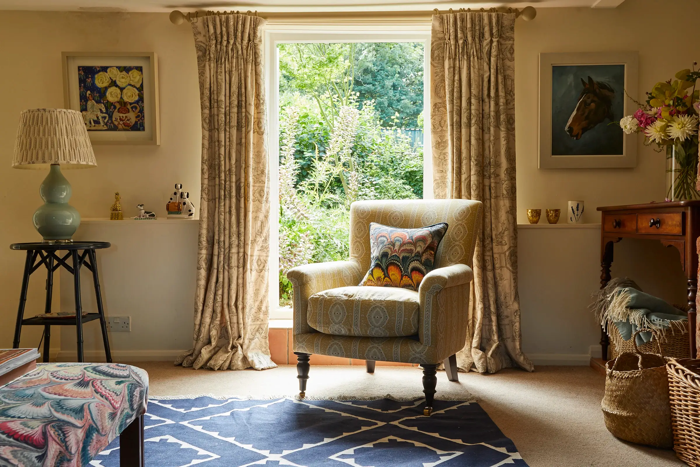 Cozy room with an armchair, patterned rug, and window view of garden. Art and a side table decorate the space.