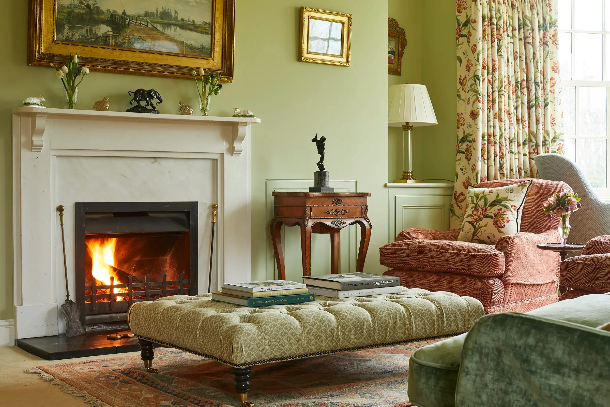 Cozy living room with a fireplace, patterned armchairs, a tufted ottoman, and decorative artworks on pale green walls.