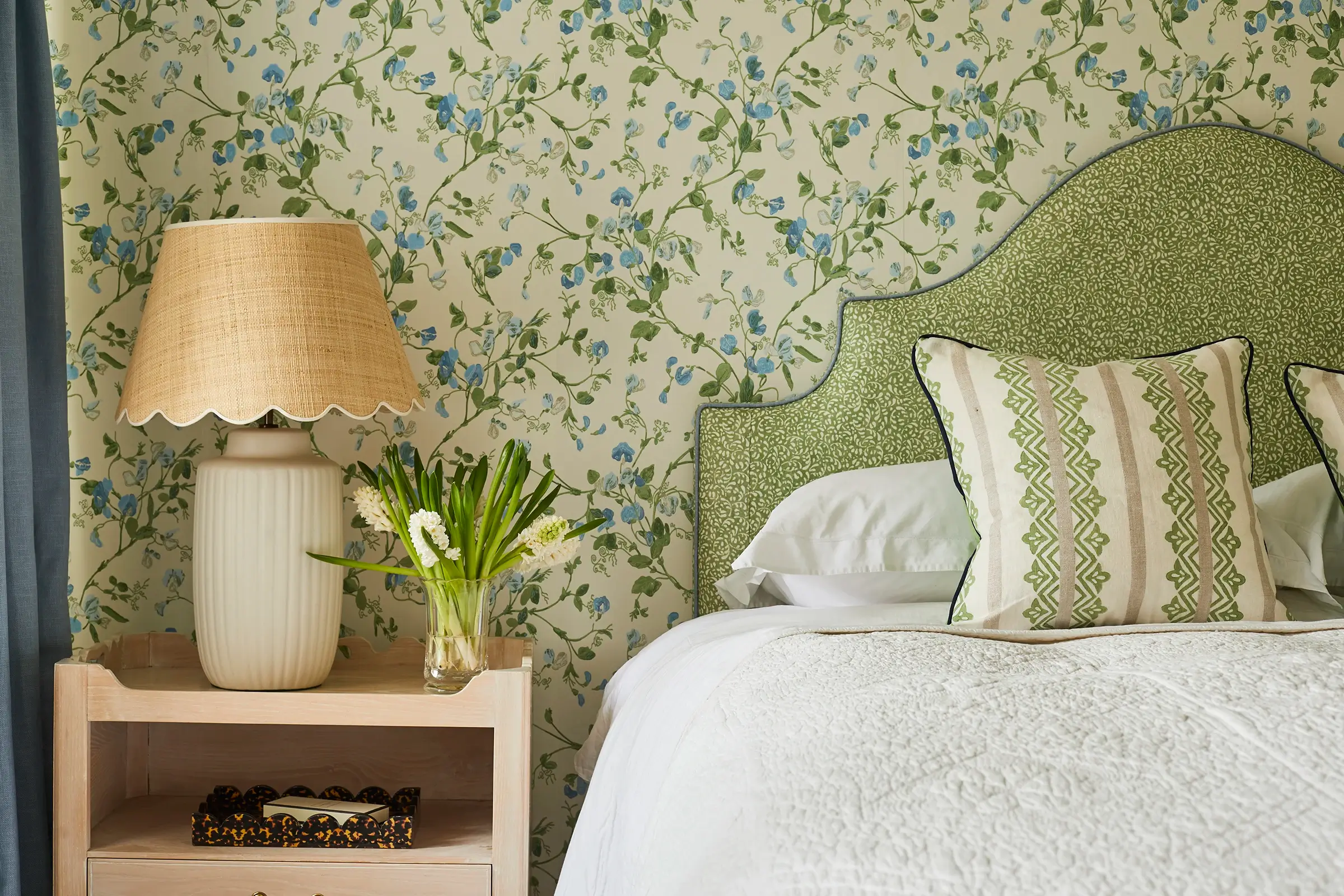 Bedroom with a floral wallpaper, green headboard, green cushion on a white bedspread with flowers and a lamp aside.