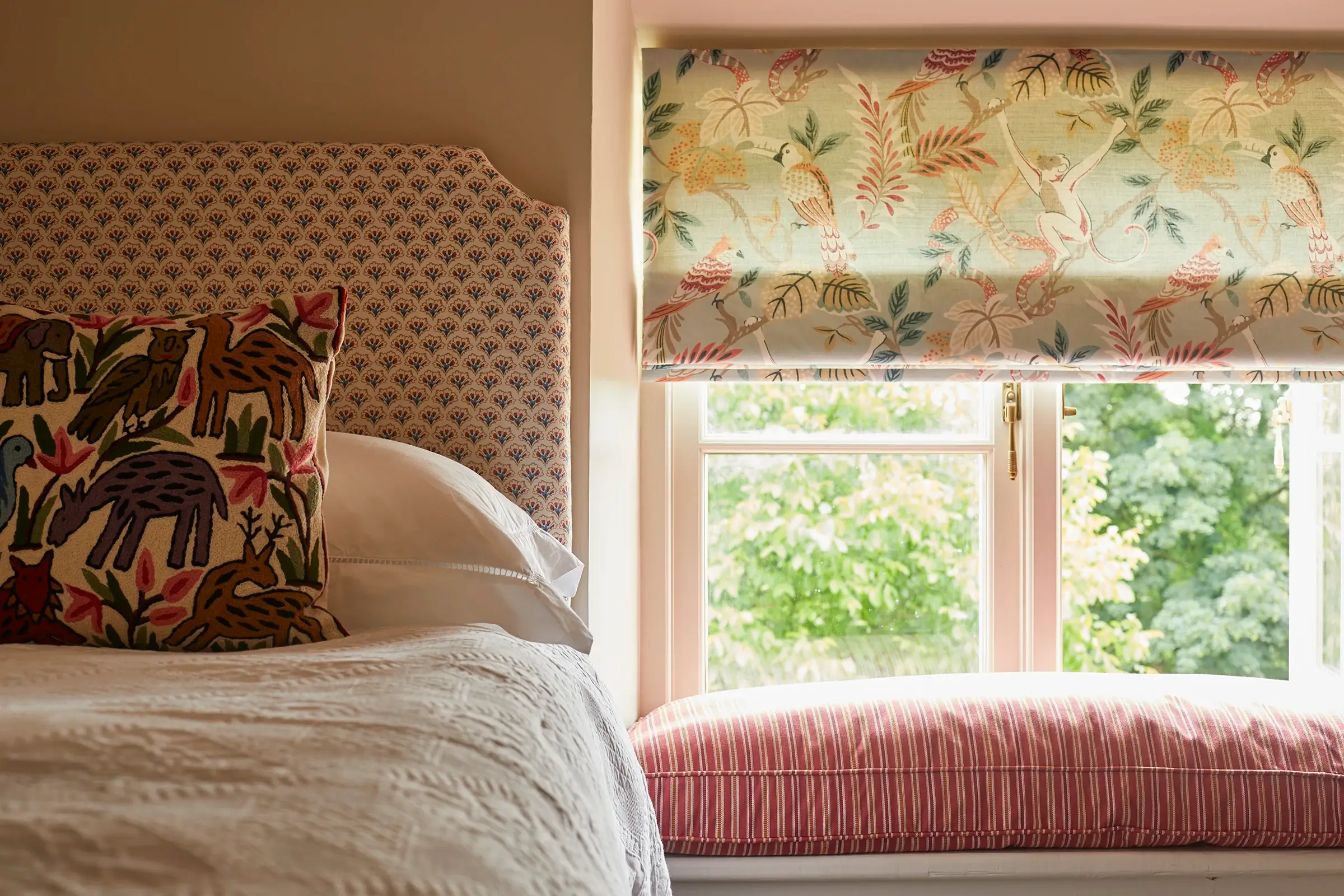 A cozy bedroom with a patterned headboard, colorful throw pillows, and a window with a floral blind overlooking greenery.