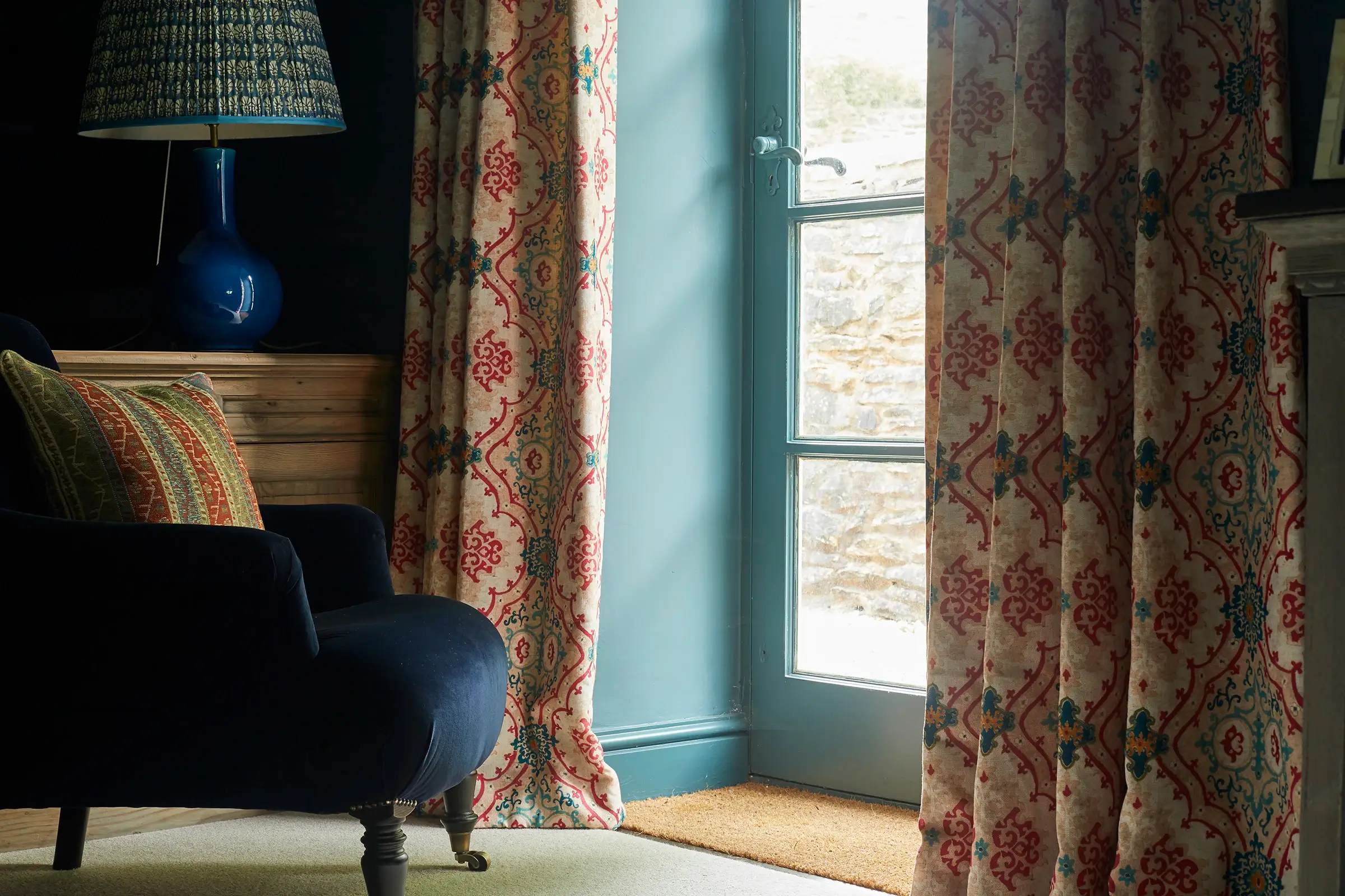 A cozy room with patterned curtains, a blue armchair, and a standing lamp near a glass door.