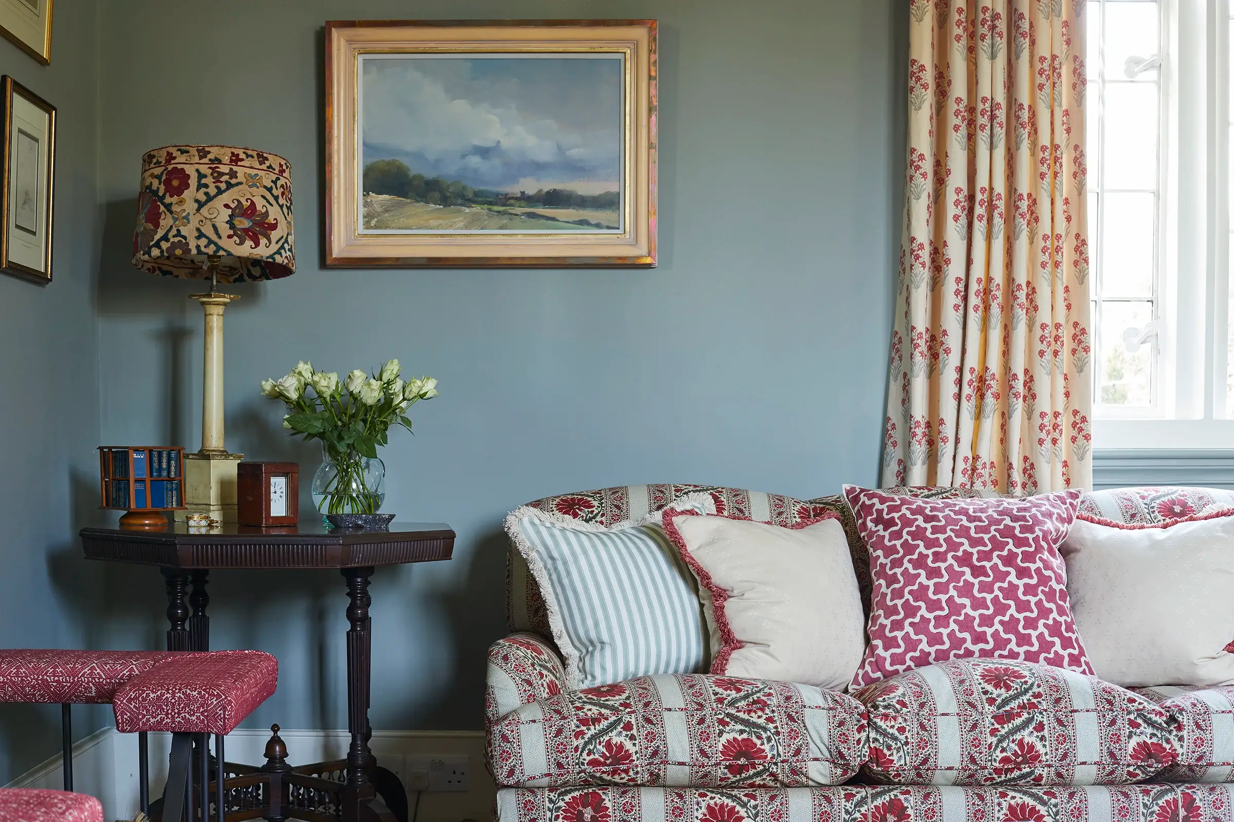 A cozy living room with a patterned sofa, pillows, flowers, and artwork on the wall.