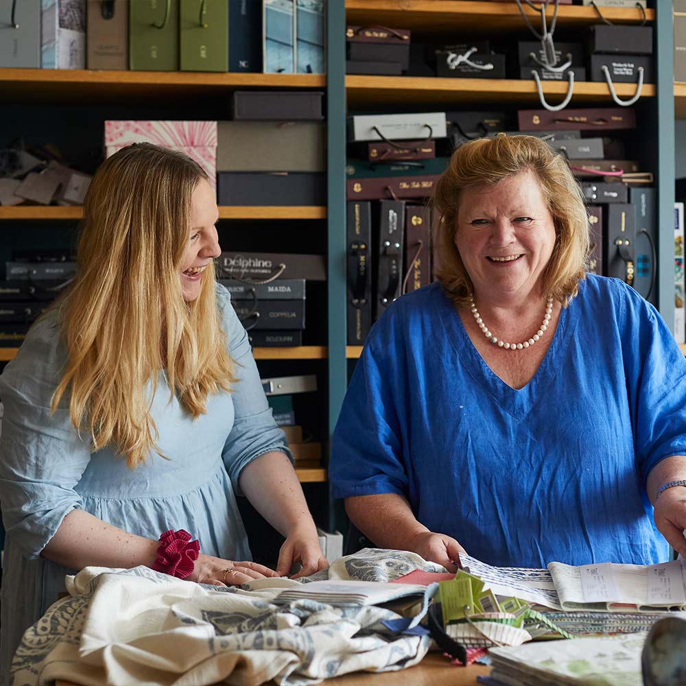 Two women smiling and discussing fabric samples in a room with shelves filled with various materials.