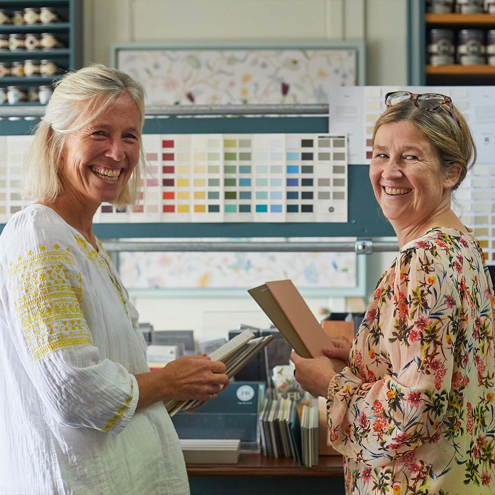 Two women smile while holding color samples in a store with paint swatches on display in the background.