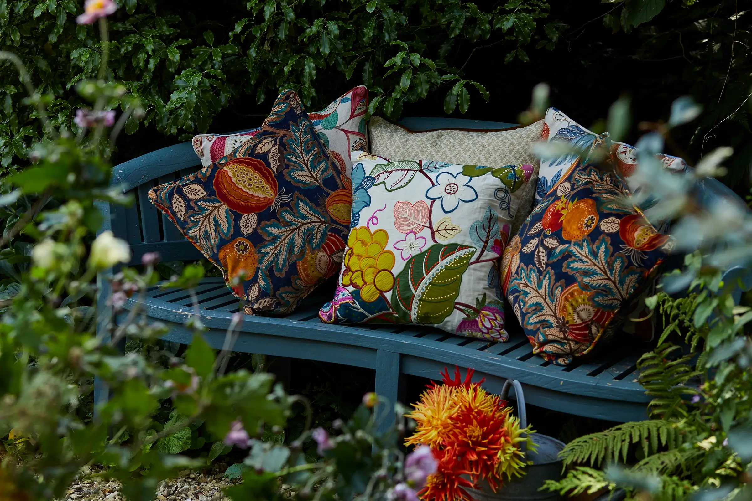 Floral-patterned pillows on a curved blue bench surrounded by greenery and flowers in a garden.