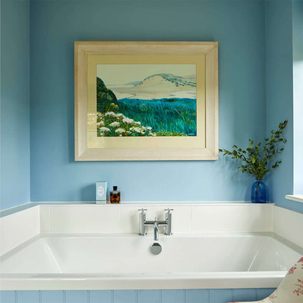 A white bathtub in a blue bathroom with a mountain painting and a vase with greenery.