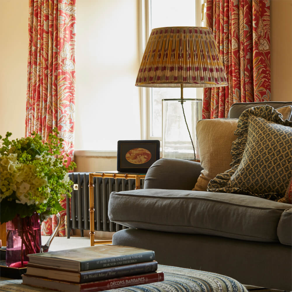 Cozy living room with floral curtains, patterned lamp, cushioned sofa, books, and a vase of flowers.