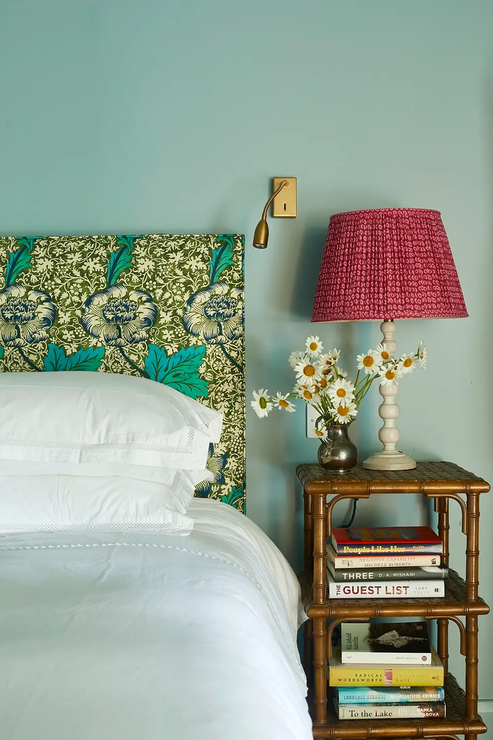 Bedroom with a floral headboard, pink lamp, daisies in a vase, and books on a wooden nightstand.