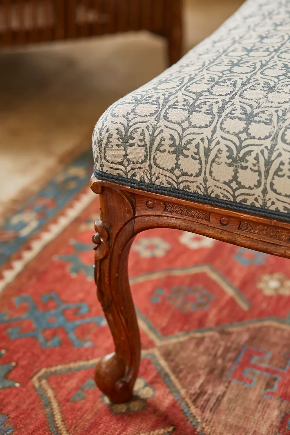 Close-up of an upholstered chair corner with a patterned cushion and ornate wooden legs on a rug.