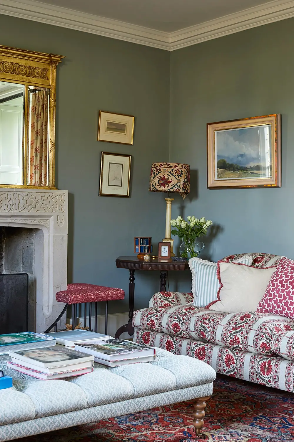 Cozy living room with patterned sofa, ottoman, framed wall art, ornate fireplace, and a floral table lamp.