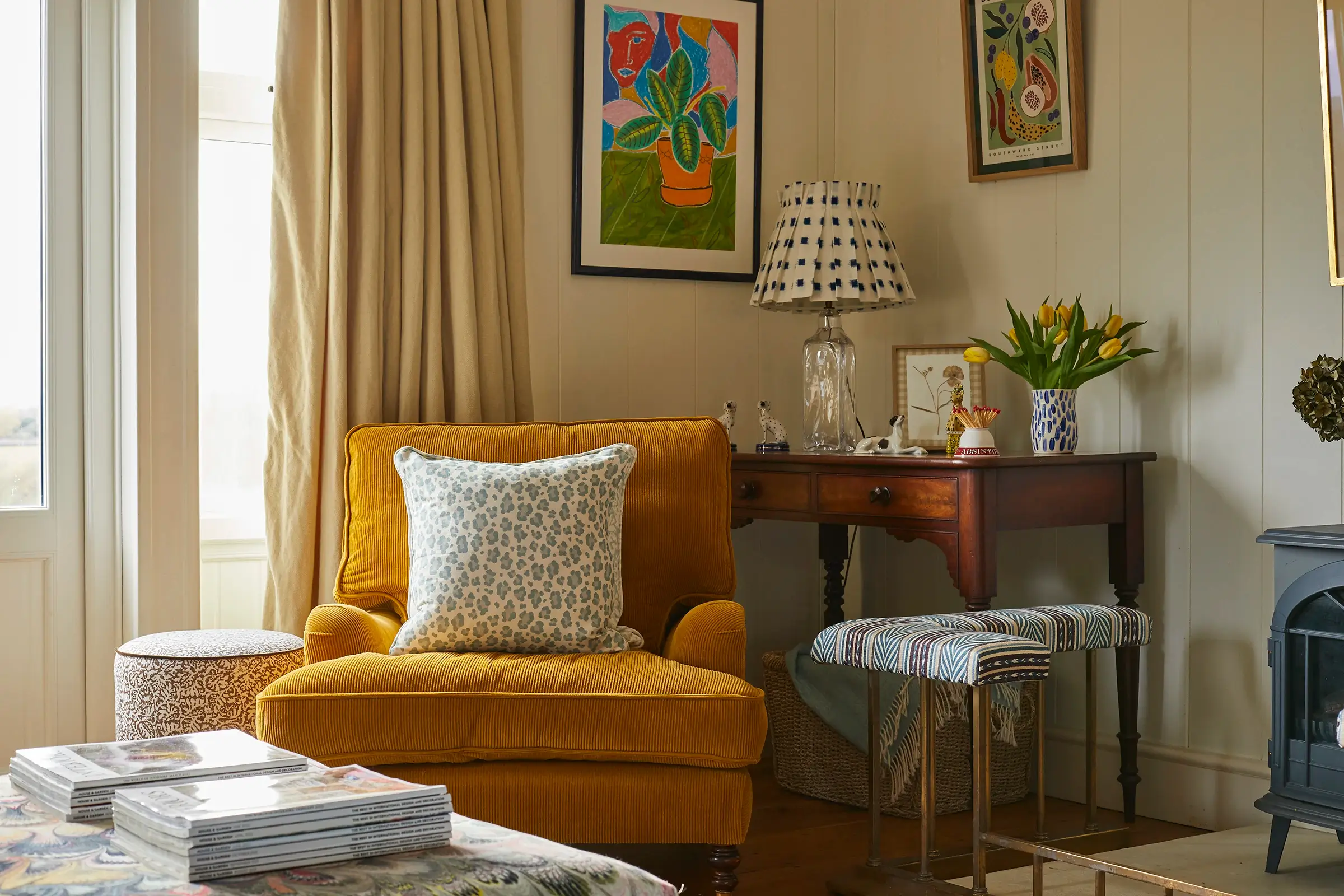 Cozy living room with a mustard armchair, floral lamp, framed art, and a wooden desk decorated with tulips.