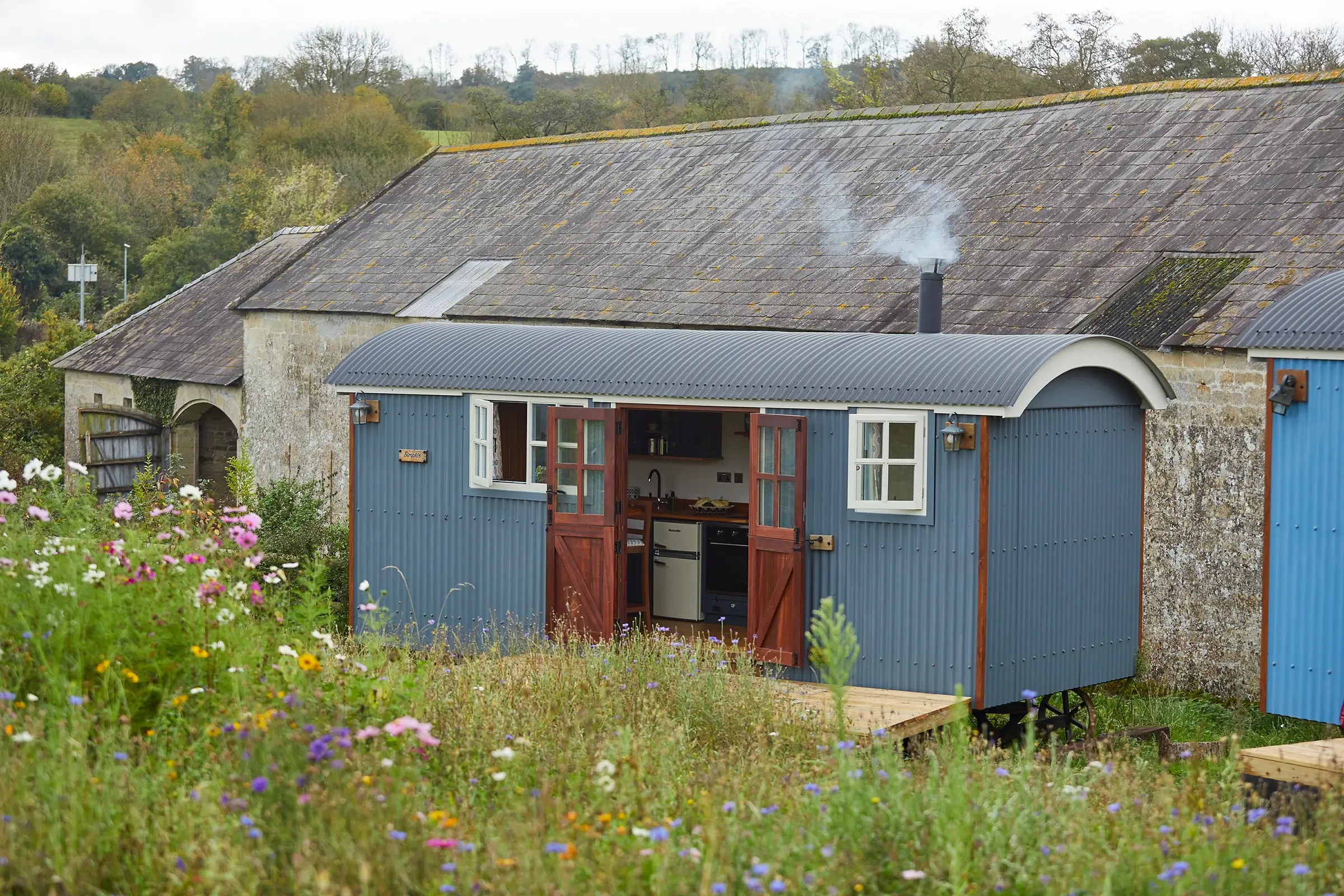 Blue shepherds hut with open doors, smoke from chimney, and wildflowers in foreground; barn and trees in background.