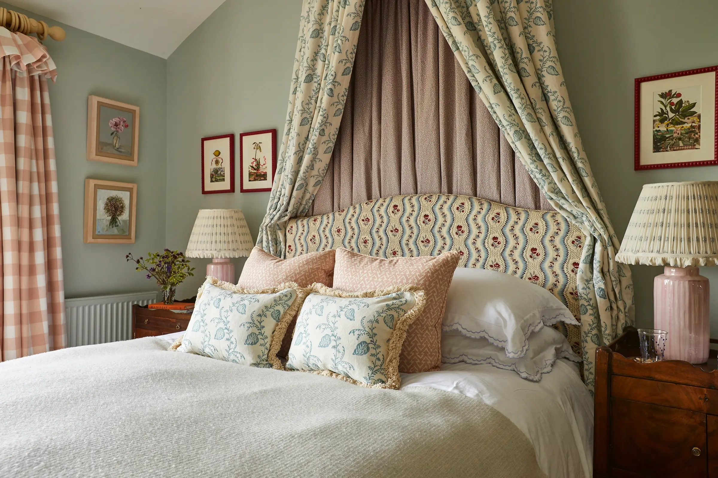 A cozy bedroom with a patterned headboard, floral curtains, framed artwork, and decorative pillows on the bed.