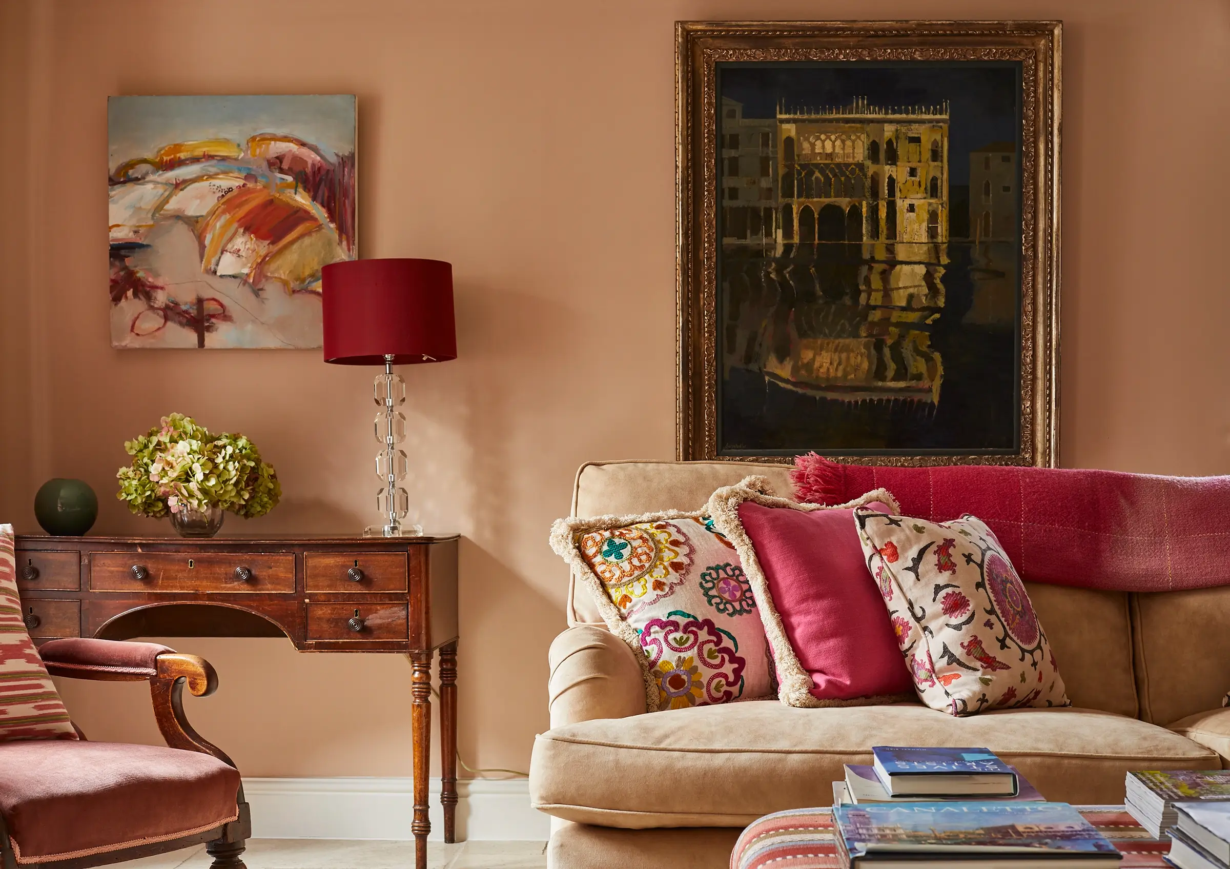 Cozy living room with a beige sofa, colorful cushions, two paintings, and a wooden table with a lamp.