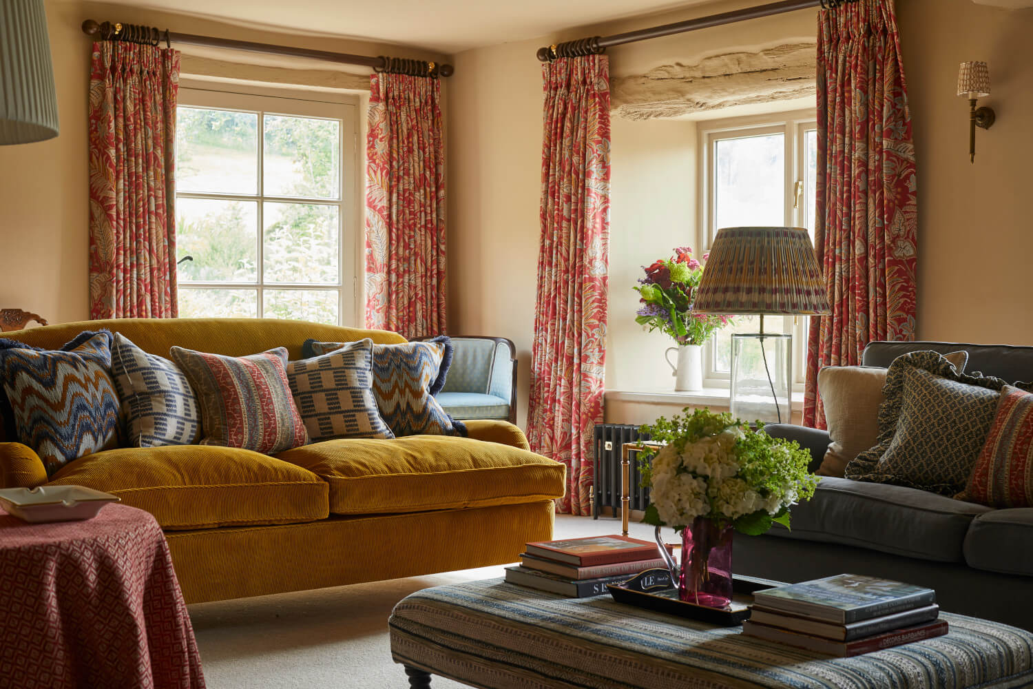 Cozy living room with colorful cushions, yellow sofa, floral curtains, and books on a table near a window with a scenic view.