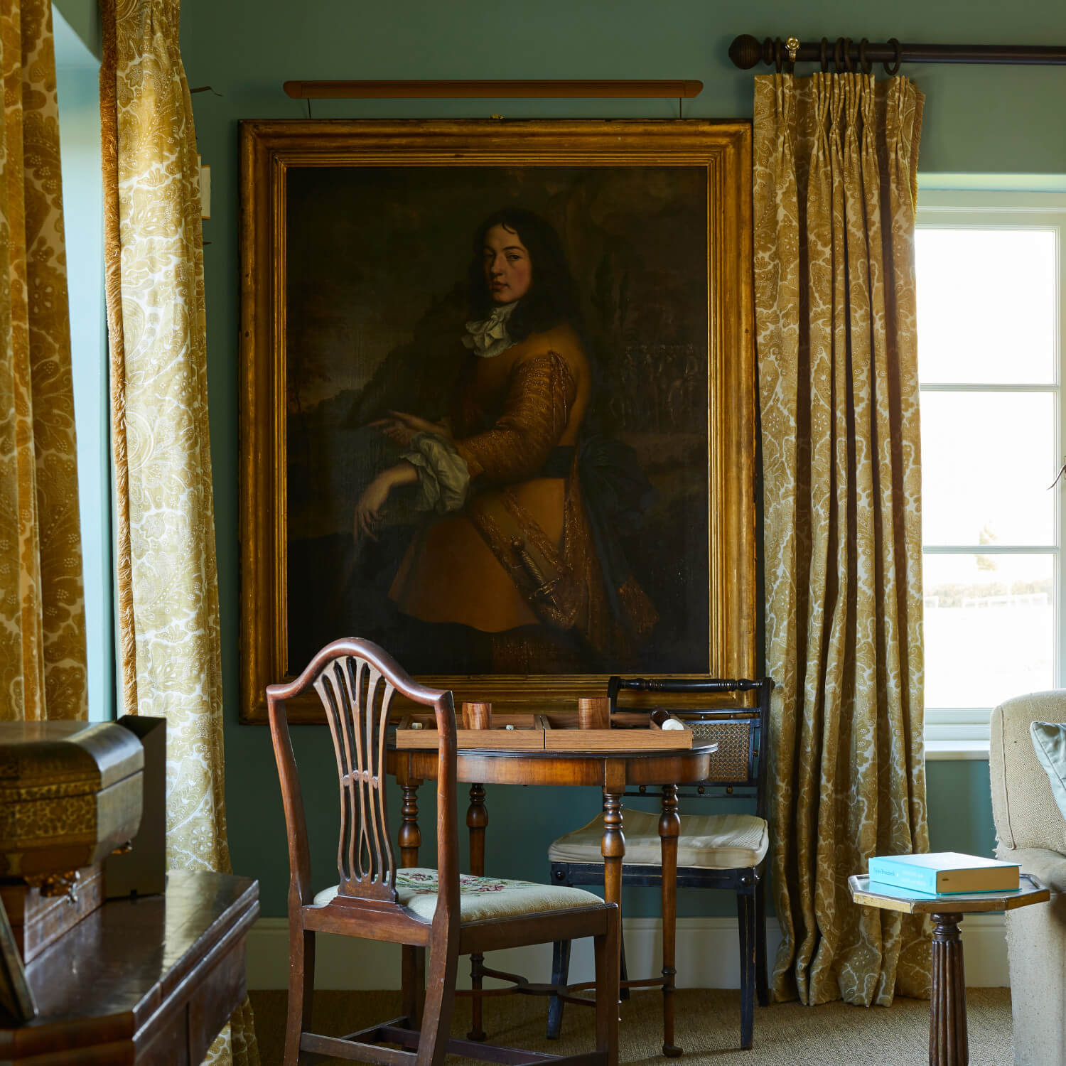 Room with a large portrait on the wall, a small wooden table, chairs, and ornate curtains by a window.