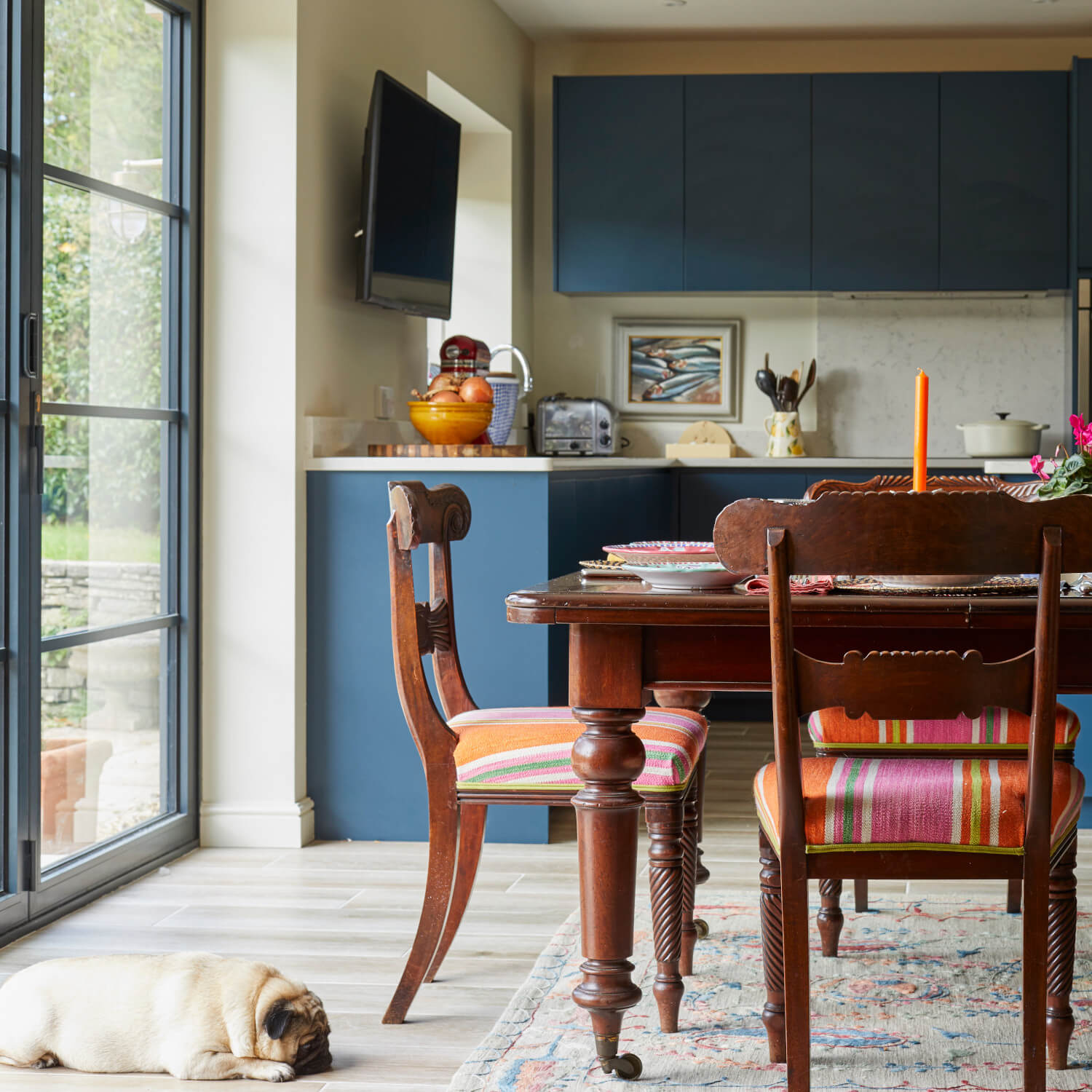 A kitchen with wooden chairs, colorful cushions, a dining table, and a pug resting on the floor.