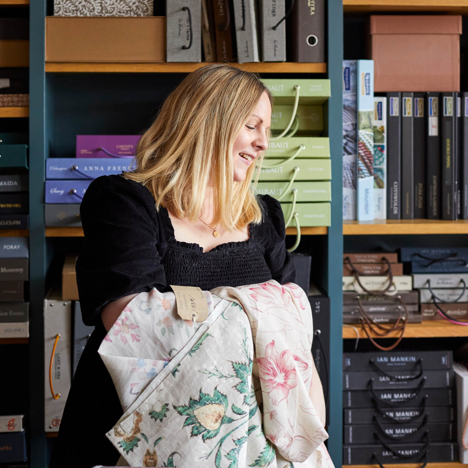 Person with blonde hair in a black dress holds patterned fabric in a shop, with shelves of branded boxes and folders in the background.